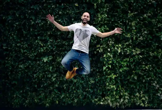 man wearing t-shirt and jeans jumpshot in front of a green hedge